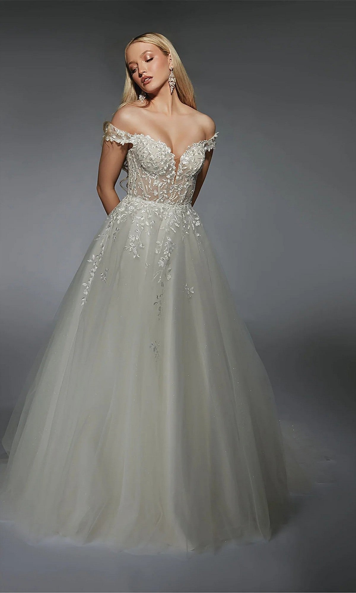 A woman wearing the Long White Dress: Alyce 7129, featuring an ivory lace corset bodice and off-the-shoulder neckline, stands against a plain gray background.